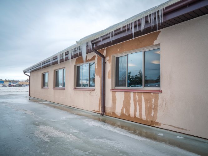 Single-story building with icicles hanging from roof and ice on ground, showing signs of water damage on walls.